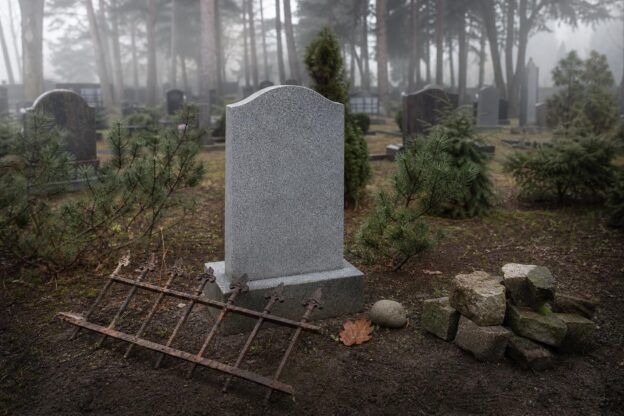 Ein grauer Grabstein auf einem Friedhof umgeben von Kiefern, mit einem verrosteten Zaunstück, einem Eichenblatt und